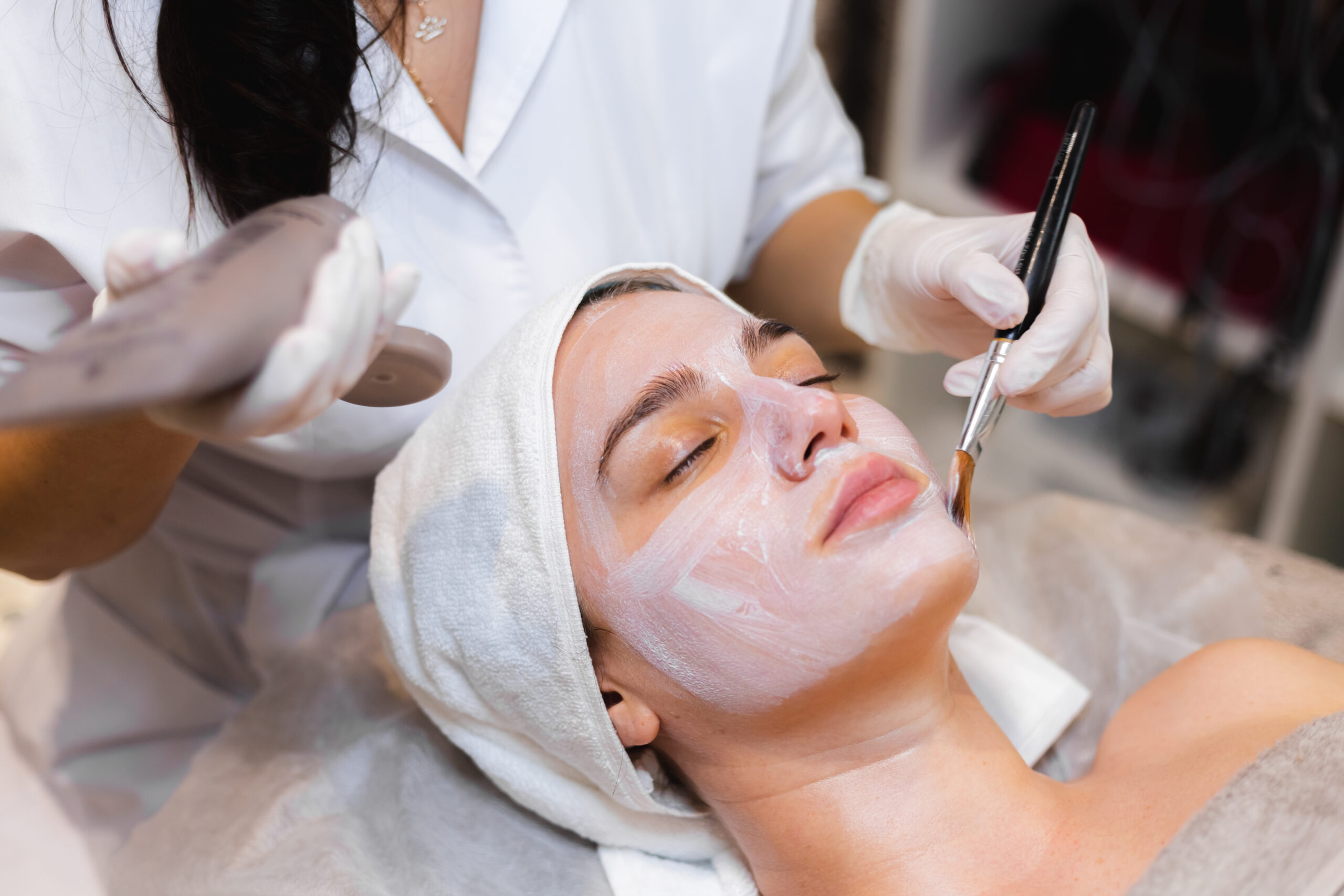beautician with a brush applies a white moisturizing mask to the face of a young girl client in a spa beauty salon